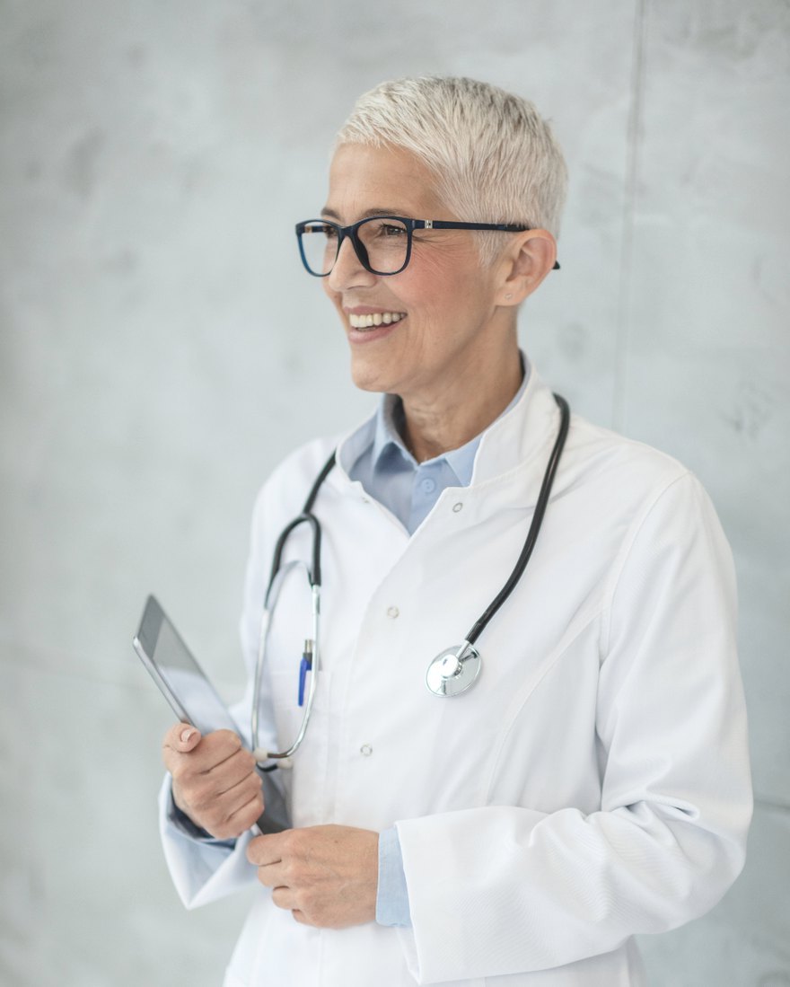 A physician in a white coat with a stethoscope, holding a tablet and smiling